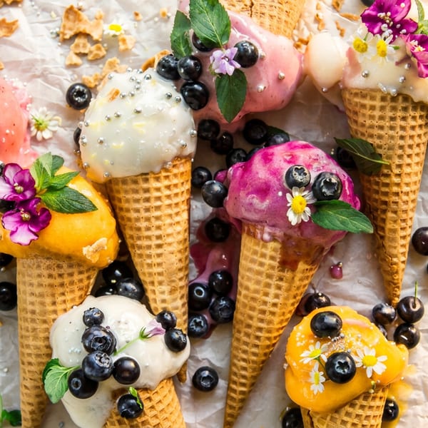 Multiple colorful ice cream cones with blueberries, sprinkles, flowers, and mint leaves on crumpled paper.