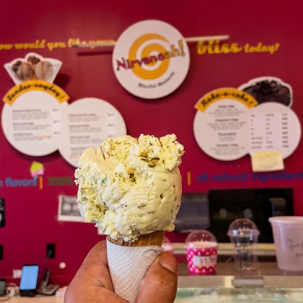 A hand holds a pistachio-flavored ice cream cone in an ice cream shop with a red wall and menus.