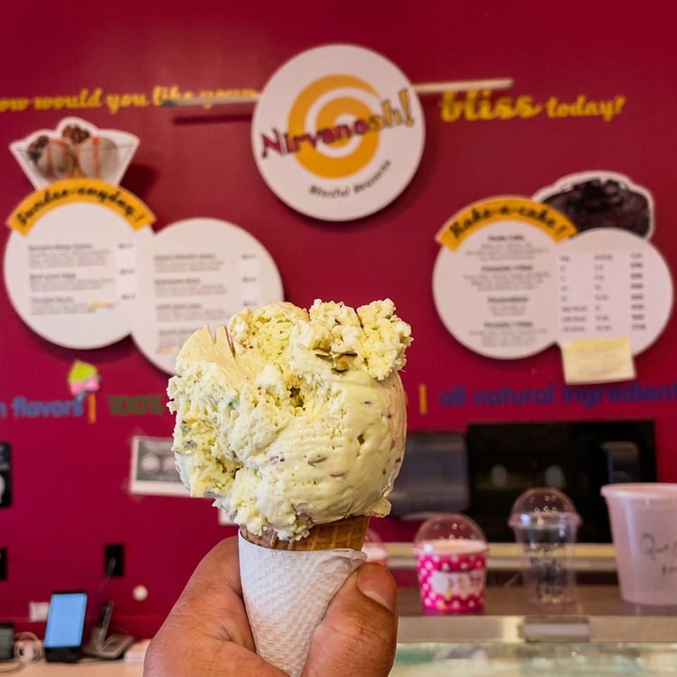 A hand holds a pistachio-flavored ice cream cone in an ice cream shop with a red wall and menus.
