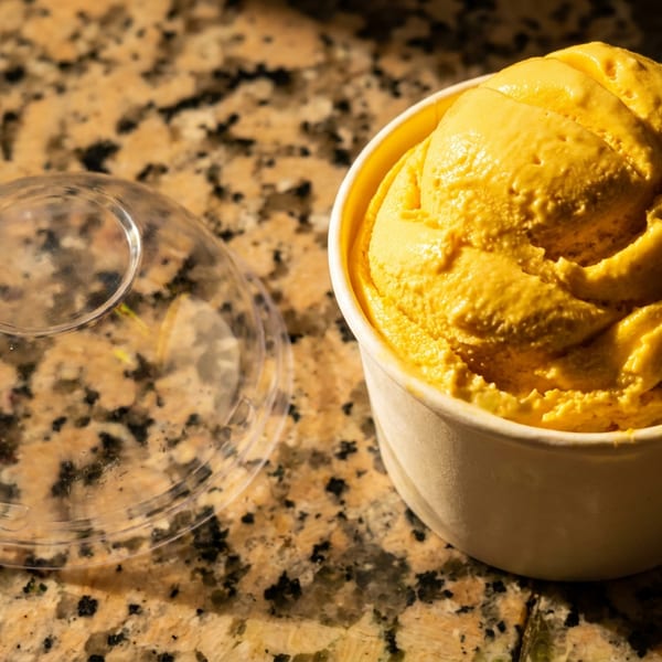 Bright yellow gelato in a white cup and a clear plastic lid on a granite countertop.