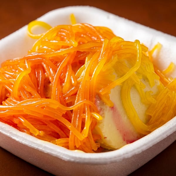 Colorful orange and yellow falooda noodles over a creamy dessert in a white takeaway container.